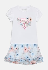 Λευκό κοντομάνικο πουκάμισο με λαμπερό λογότυπο "GUESS" και λουλουδάτο μοτίβο με πεταλούδες. Συνδυασμένο με μια ανοιχτό μπλε, βολάν φούστα με λουλουδάτο σχέδιο.