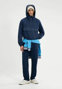 Dunkelblaues Pullover-Jacke mit Kapuze und Reißverschluss, kombiniert mit dunkelblauen Jogginghosen. Blauer Pullover um die Taille gebunden und weiße Sneakers mit schwarzen Akzenten.