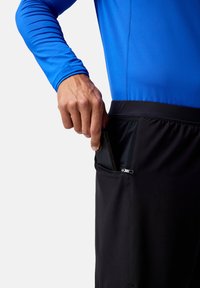 Pantalones cortos deportivos negros con un bolsillo con cremallera en el lateral. Se lleva puesto una camiseta de manga larga azul. Un smartphone está parcialmente insertado en el bolsillo.