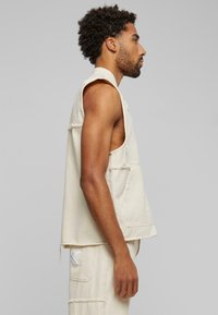 Beige ermeløs vest laget av strukturert stoff, med sidelommer og slitte kanter for en avslappet passform.