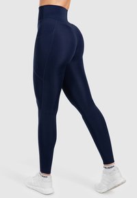 Leggings taille haute en marine, fabriquées dans un tissu extensible, avec des poches latérales et un design épuré, associées à des chaussures de sport blanches.