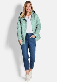 Gequiltede mintgroene jas met een capuchon, gecombineerd met een witte top en blauwe jeans. Heeft drukknoopsluitingen en een relaxte pasvorm.
