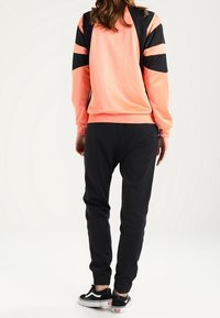 Korálová mikina s černými doplňky, s kulatým výstřihem a pruhovanými rukávy; doplněná černými zúženými tepláky a černými teniskami.