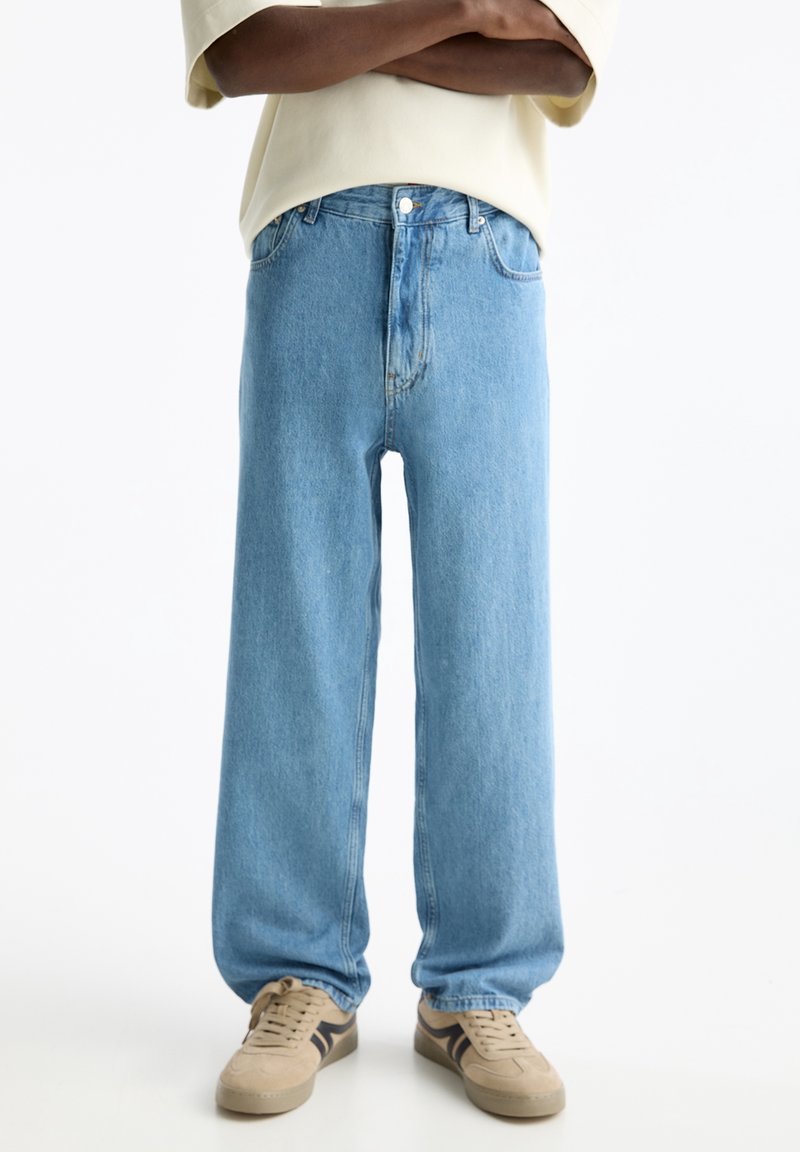 Personne portant un jean large bleu clair et des baskets beige, debout les bras croisés devant un fond blanc uni.