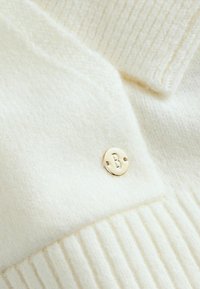 Krēmgadīgā adītā džemperis ar ribotu apkakli un aprocēm, ar zelta apaļu logo birku, uz kuras ir "B". Redzama mīksta tekstūra.