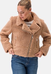 Tansfarbige Kunstfelljacke mit Reverskragen, silbernem Reißverschluss, seitlichen Taschen und strukturiertem Stoff. Tragen über einem weißen Oberteil und blauen Jeans.
