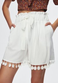 White shorts with a tie waist and elastic band, featuring side pockets and a decorative hem with round trim accents. Smooth fabric texture.