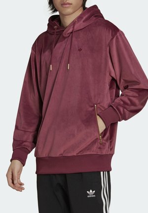 Junge Person trägt einen weinroten Velours-Hoodie mit goldener Reißverschlusstasche und schwarze Adidas-Hosen mit weißen Streifen und Logo.