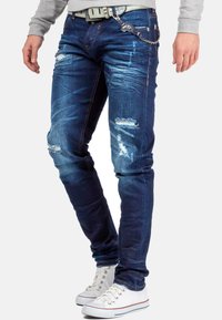 Jeans in denim blu con una vestibilità slim, caratterizzati da toppe strappate e un dettaglio a catena sul passante della cintura. Completi di tasche tradizionali.