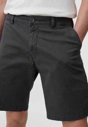 Homme portant un short gris foncé boutonné et une chemise blanche, les mains dans les poches, cadré à la taille et aux genoux.