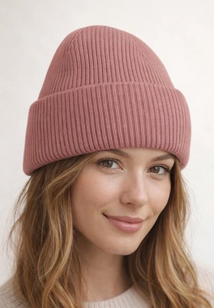 Young woman with light brown hair wearing a pink ribbed knit beanie, smiling softly against a plain light background.