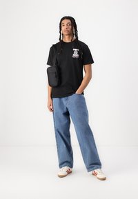 Svart T-skjorte med grafisk trykk, blå baggy jeans og hvite joggesko med røde striper. Svart crossover-veske henger over skulderen.