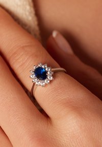 Saffierring met een ronde blauwe steen omringd door kleinere heldere stenen op een zilveren band, met een bloemmotief en een gepolijste afwerking.