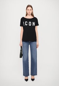 T-shirt noir à manches courtes avec un grand lettrage blanc "ICON", associé à un jean bleu à jambes larges et à des escarpins noirs à bout pointu. Tenant un sac à main noir.