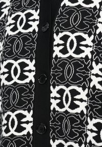 Melnā un baltā raksta džemperis ar adījuma tekstūru, apaļām melnām pogām un simetrisku dizainu uz auduma.