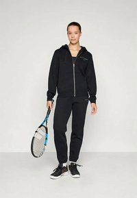 Melnā zip-up kapuce ar šņorēm, apvienota ar atbilstošām melnām sporta biksēm. Modelis tur zilas un baltas tenisa raketes, ģērbts melnos sporta apavos.