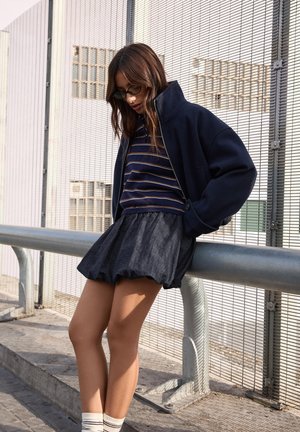 Jeune femme portant des lunettes, un haut rayé, une veste marine et une jupe bouffante foncée, appuyée contre une rampe métallique près d'une clôture en fil de fer à l'extérieur.