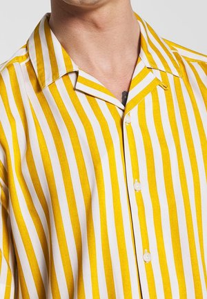 Man draagt een geel-wit verticaal gestreept overhemd met kraag en knopen, waarbij een deel van zijn nek en bovenborst zichtbaar is.