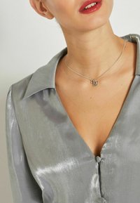 Gray satin shirt with a collared neckline, button closures, and a subtle sheen. Silver necklace featuring an "M" pendant.