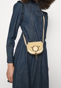 Borsa a tracolla in pelle beige chiaro con pattina arrotondata, anello in hardware dorato e borchie decorative. Include un portachiavi staccabile.