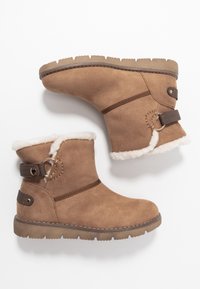 Bottines en suede tan avec bord en fausse fourrure blanche, présentant une sangle marron et un accent de matériel rond. Semelle en caoutchouc épais avec un motif texturé.