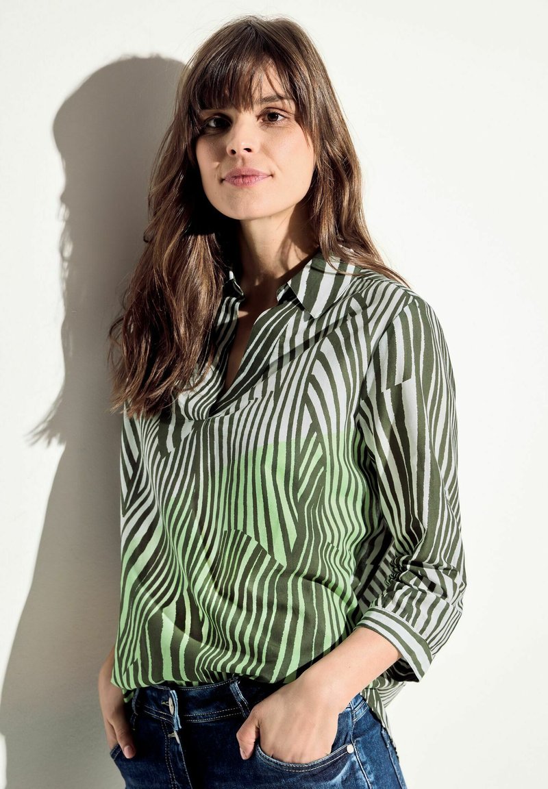 Blusa a righe verdi e bianche con collo e maniche a tre quarti, abbinata a jeans in denim scuro. Tessuto morbido.
