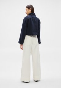 Femeie purtând o jachetă scurtă bleumarin și pantaloni albi cu picioare largi, stând în picioare și privindu-și spre stânga, pe un fundal simplu, deschis la culoare.