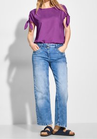 Blouse violette à manches courtes avec des manches attachées, associée à un jean large bleu clair et des sandales navy à boucles. Fond clair.