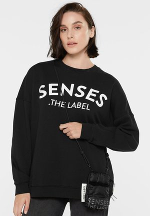 Schwarzes oversized Sweatshirt mit weißem Text "SENSES .THE LABEL" auf der Vorderseite. Verfügt über einen runden Ausschnitt und lange Ärmel. Hält eine kleine schwarze Tasche.