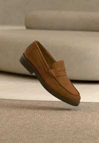 Mocassin en daim marron avec semelle foncée flottant au-dessus d'un tapis beige, canapé beige en arrière-plan.