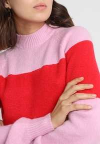 Femme portant un pull rose avec une large bande rouge sur la poitrine, les bras croisés, montrant un teint naturel et un maquillage léger.