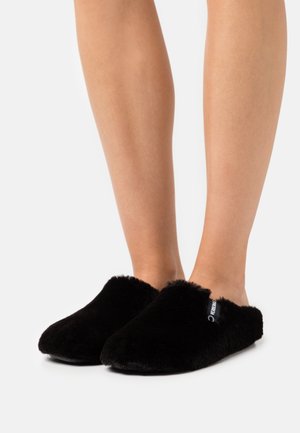 Schwarze, flauschige Hausschuhe mit runder Zehenpartie, weichem Material und einem Seitenschild. Das Design bietet einen schlupfbaren Stil für einfaches Anziehen.