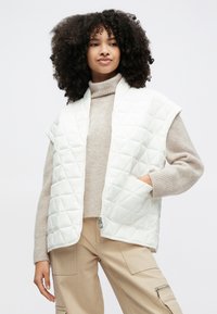 Balta dygsniuota liemenė su trumpomis rankovėmis, užsegama užtrauktuku ir turi šonines kišenes. Dėvima ant šviesiai rudos aukšta apykakle megztinio ir smėlio spalvos kargo kelnių.