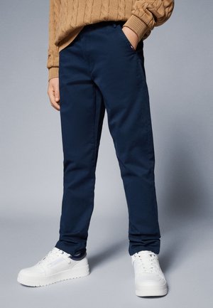 Person iført marineblå bukser, hvide sneakers og en lysebrun kabelstrikket sweater med den ene hånd i lommen mod en grå baggrund.
