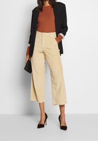 Beige corduroy leveälahkeiset housut korkealla vyötäröllä. Yhdistetty ruskean riipustetun paidan ja mustan bleiserin kanssa, lisävarusteena musta clutch-laukku.