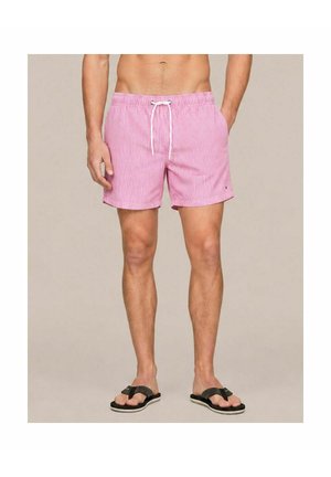 Man wearing pink and white striped swim shorts with white drawstring and black flip-flops standing against a plain background.