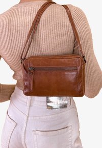 Hnědá kožená crossbody taška s přední kapsou na zip, obdélníkového tvaru a tenkými ramenními popruhy, nošená přes texturovaný béžový top.