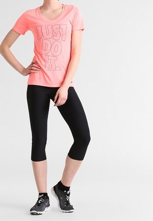 Kvinde iført koralfarvet "JUST DO IT." t-shirt, sorte cropped leggings og sorte sneakers, stående med hånden på hoften.