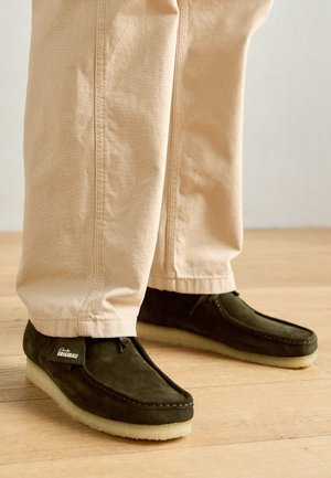 Grüne Wildleder-Mokassins mit einer Schnürung und einer cremefarbenen, strukturierten Gummisohle, kombiniert mit hellbeigen Hosen.