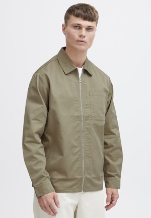 Ung mand iført en lysebrun lynlåstrøje med brystlomme og hvide bukser, stående foran en ensfarvet hvid baggrund.