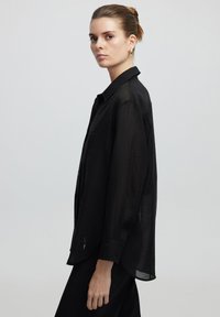 Camicia nera trasparente con bottoni e maniche lunghe, caratterizzata da una texture sottile e un orlo curvo. Il modello è in piedi contro uno sfondo neutro.
