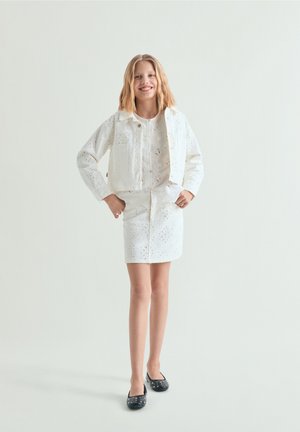 Nuori tyttö hymyilee, hänellä on olkapäille ulottuvat vaaleat hiukset, valkoinen pitsipintainen takki ja hame, sekä mustat niitit sisältävät balleriinat.