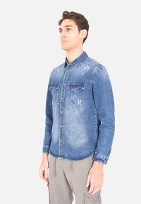 Camicia in denim azzurro chiaro con colletto classico, due tasche sul petto e dettagli vissuti. Maniche lunghe con polsini abbottonati e cuciture laterali.