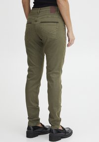 Olijfgroene broek met een taps toelopende pasvorm, zij- en achterzakken en een gestructureerde tailleband. Gecombineerd met glanzende zwarte schoenen.