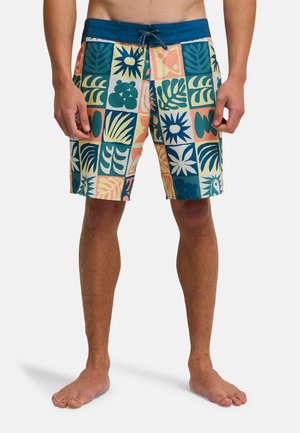 Mężczyzna w kolorowych szortach plażowych z wielokolorowymi kwadratowymi wzorami zawierającymi liście, rośliny i abstrakcyjne kształty, stojący boso na białym tle.