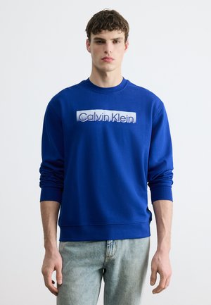 Νεαρός άνδρας με μπλε φούτερ Calvin Klein και ανοιχτόχρωμο τζιν, που στέκεται μπροστά σε απλό λευκό φόντο.