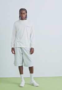 Chemise blanche à manches longues avec un petit logo, associée à un short gris clair et des baskets blanches. Design simple, coupe décontractée et texture lisse.