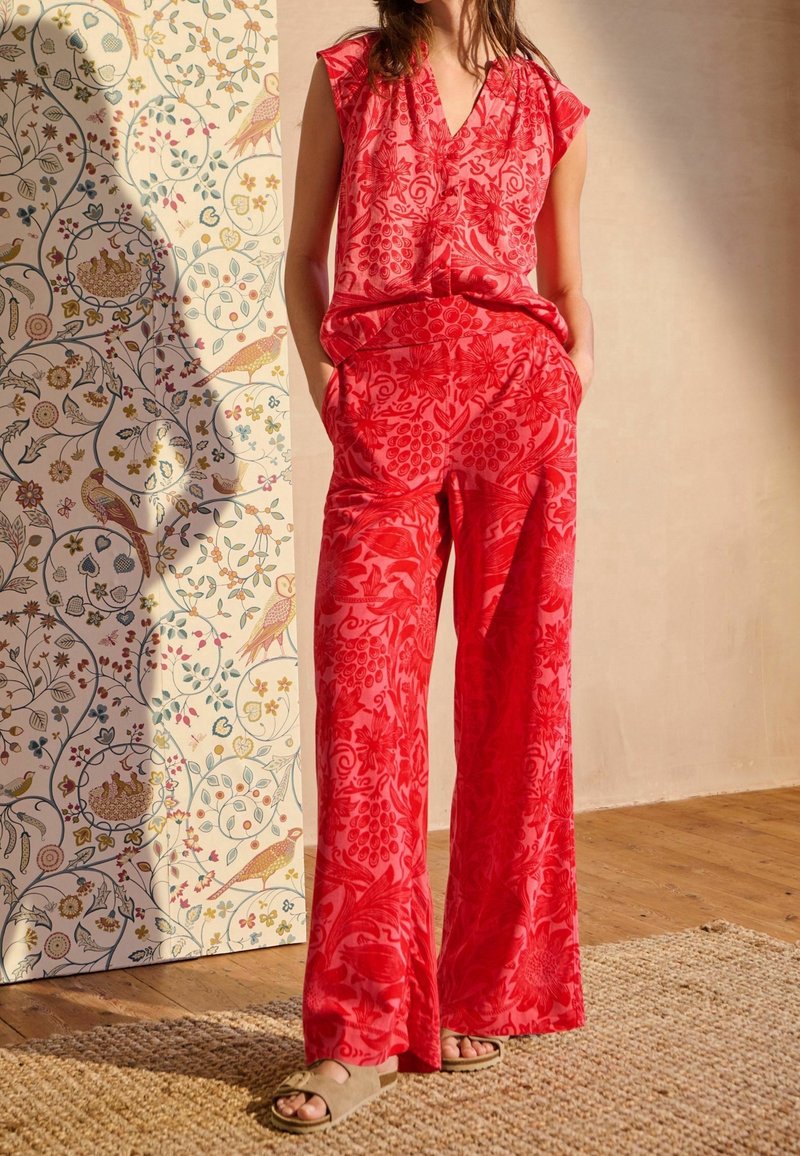 Femme debout à l'intérieur portant un haut sans manches rouge à motif floral et un pantalon large assorti, sandales beige, à côté d'un panneau mural décoratif floral.