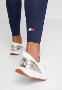 Námořnické sportovní legíny s malým červeným, bílým a modrým logem u kotníku, spárované s bílými teniskami s metalicky stříbrnou patou.