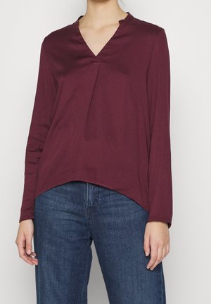 Vrouw die een lange mouwen, bordeauxrode V-hals blouse draagt, losjes ingestopt in blauwe spijkerbroek, staand tegen een effen achtergrond.
